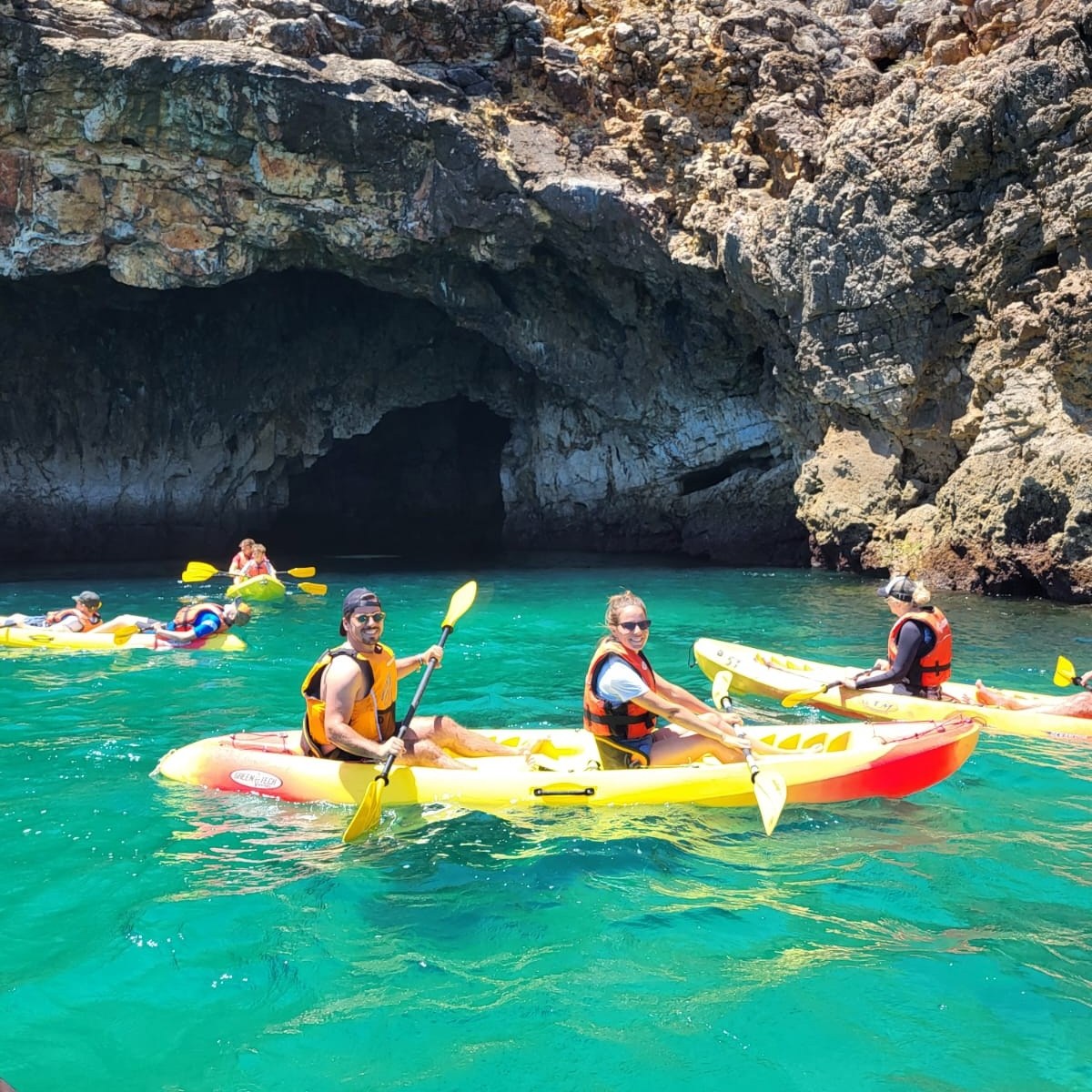 People kayaking near rocky cave entrance in clear turquoise water.