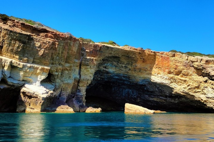 Rocky coastal cliffs with caves and clear blue water under a blue sky.