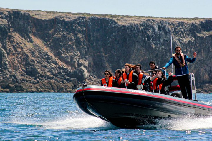 Group on a speedboat wearing life vests near a rocky shoreline.