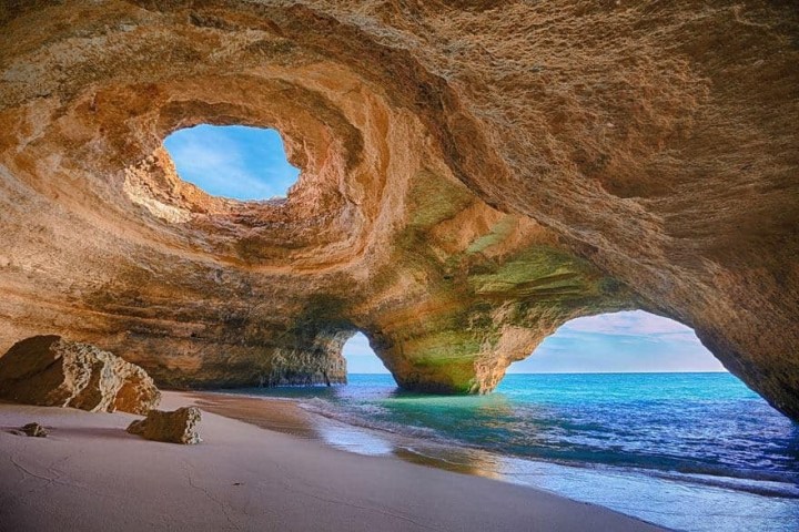 Cave with two large openings overlooking a sandy beach and ocean.