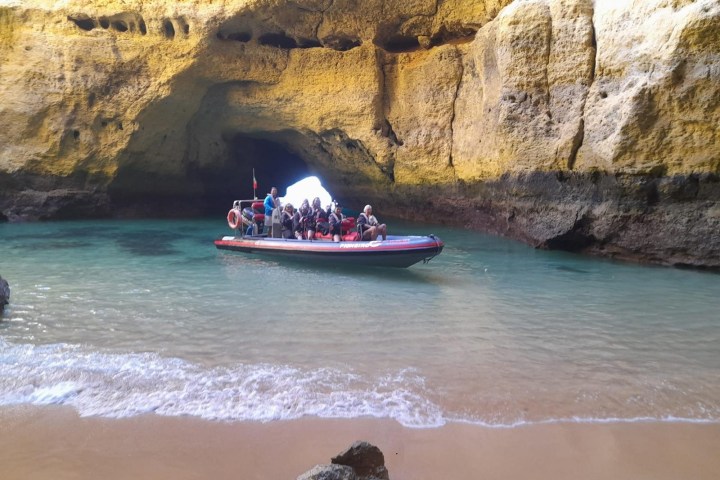 Boat with people entering a rocky cave from a sandy beach.