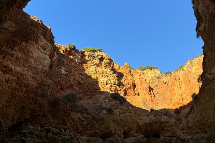 Cave opening to a beach with clear blue sky and sunlit cliffs.