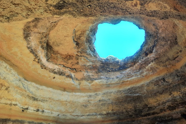 View of bright blue sky through a circular cave opening in rocky ceiling.